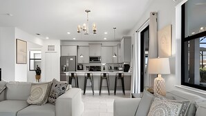 Een moderne keuken met witte kasten, een centraal eiland en barstoelen. Er zijn twee witte banken met decoratieve kussens.