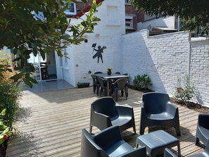 Un patio al aire libre con una terraza de madera, una mesa con sillas y una pared de ladrillo blanco con un mural en silueta.