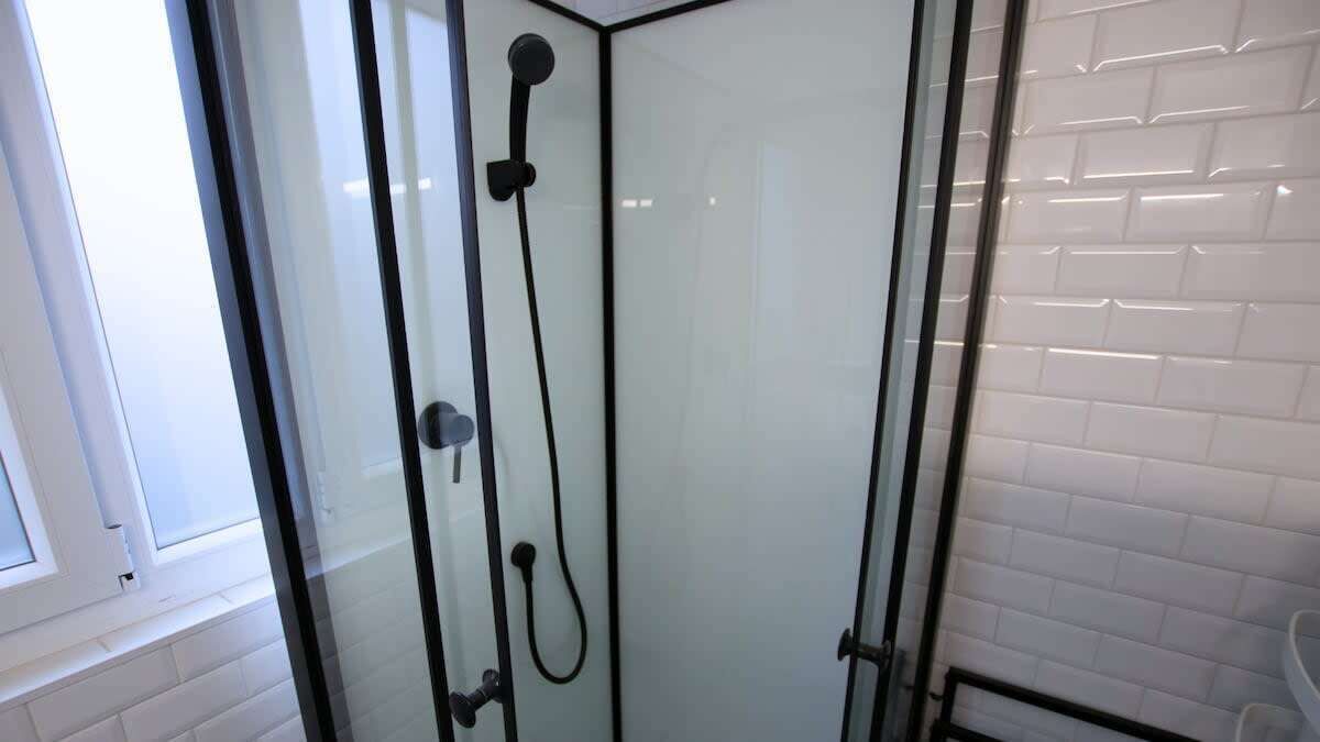 A glass-enclosed shower with a black frame, white tiled walls, and a window to the left.