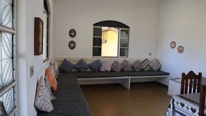 Een kamer met een ingebouwde bank langs een muur, een raam met een open luik, en een stoel met een tafelkleed met patroon.