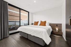 A modern bedroom with a large bed, a nightstand, a bedside table, and a view through a sliding glass door. - Sophisticated Inner-city 2-bed With Outdoor Dining (Canberra)
