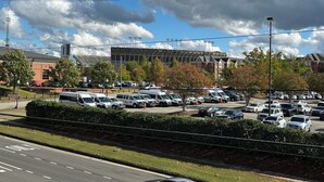 A parking lot with multiple vehicles, a road with a "No Entry" sign, and a large building with a stadium in the background.