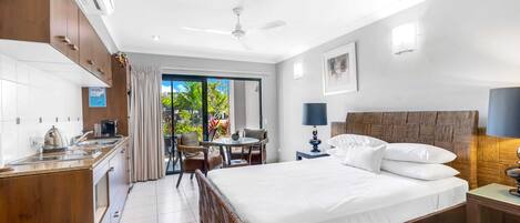Habitación de hotel con una cama grande, un escritorio con silla, una pequeña mesa con sillas, un ventilador de techo y vistas a zonas verdes.