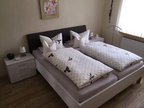 Uma cama de casal com roupa de cama florida, uma mesa de cabeceira com abajur, um criado-mudo com relógio e uma obra de arte pendurada na parede.