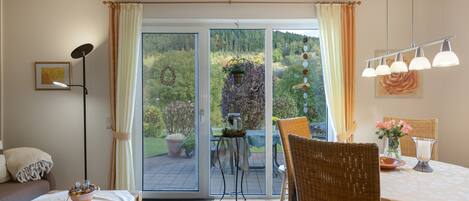 A room with a dining table, chairs, a sofa, a floor lamp, a window with curtains, and a view of the outdoors.