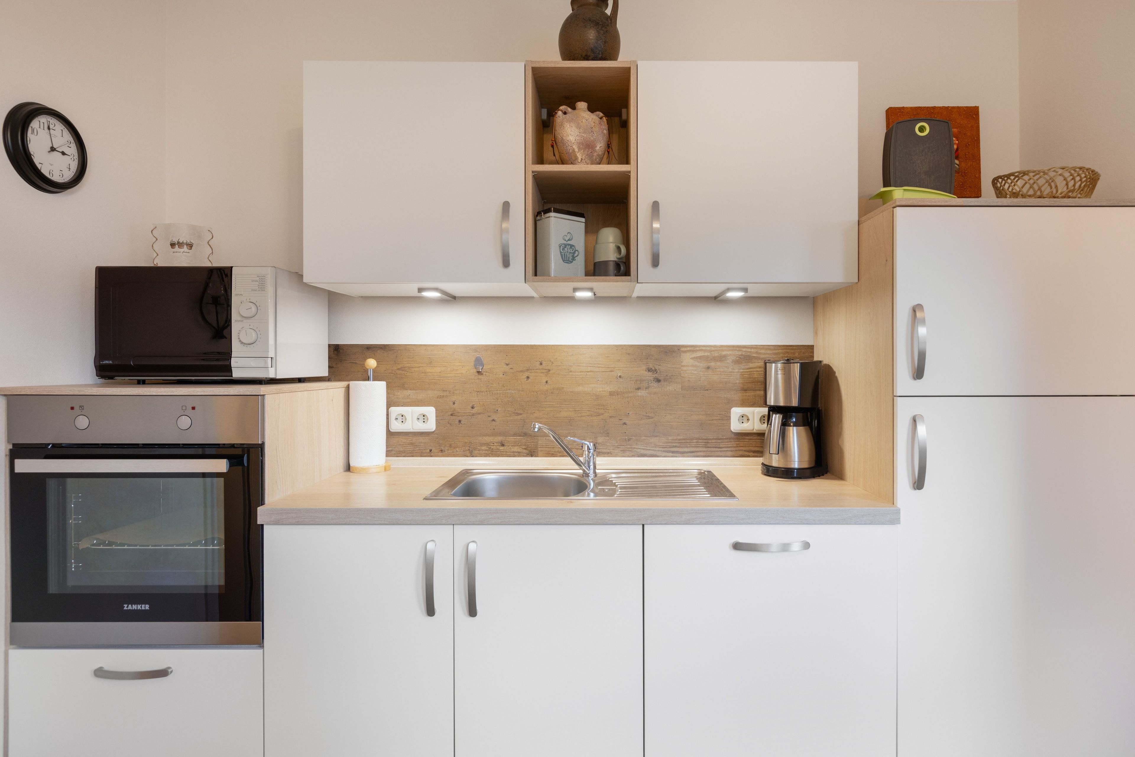 Una cocina moderna con armarios blancos, horno empotrado, cafetera y un reloj en la pared.
