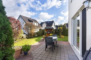 Un espace patio aménagé avec une table et des chaises, entouré de plantes en pot et d’une pelouse bien entretenue.
