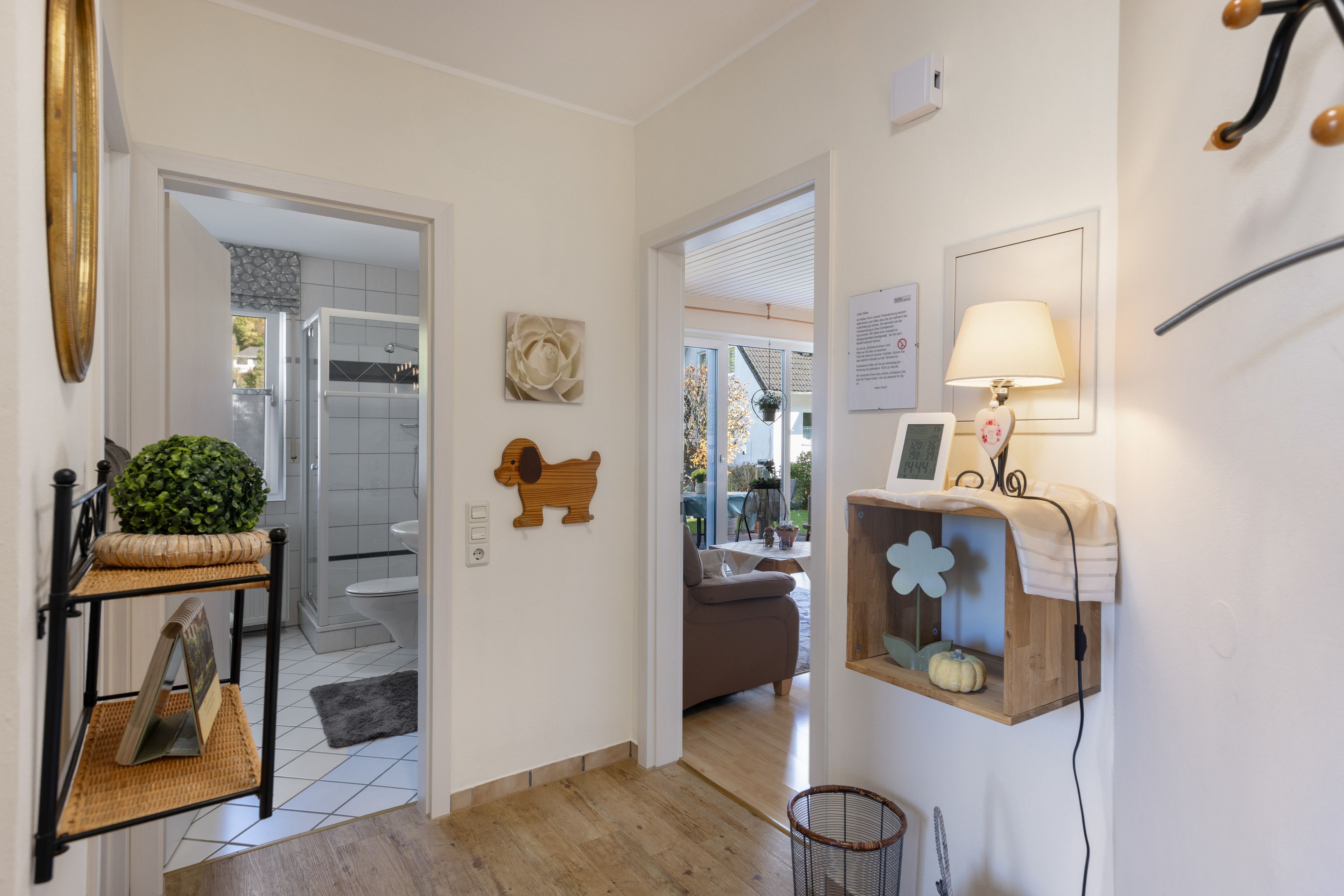 A hallway with a wooden floor, a mirror, a small plant, a dog figurine, a wall-mounted lamp, and a framed picture.