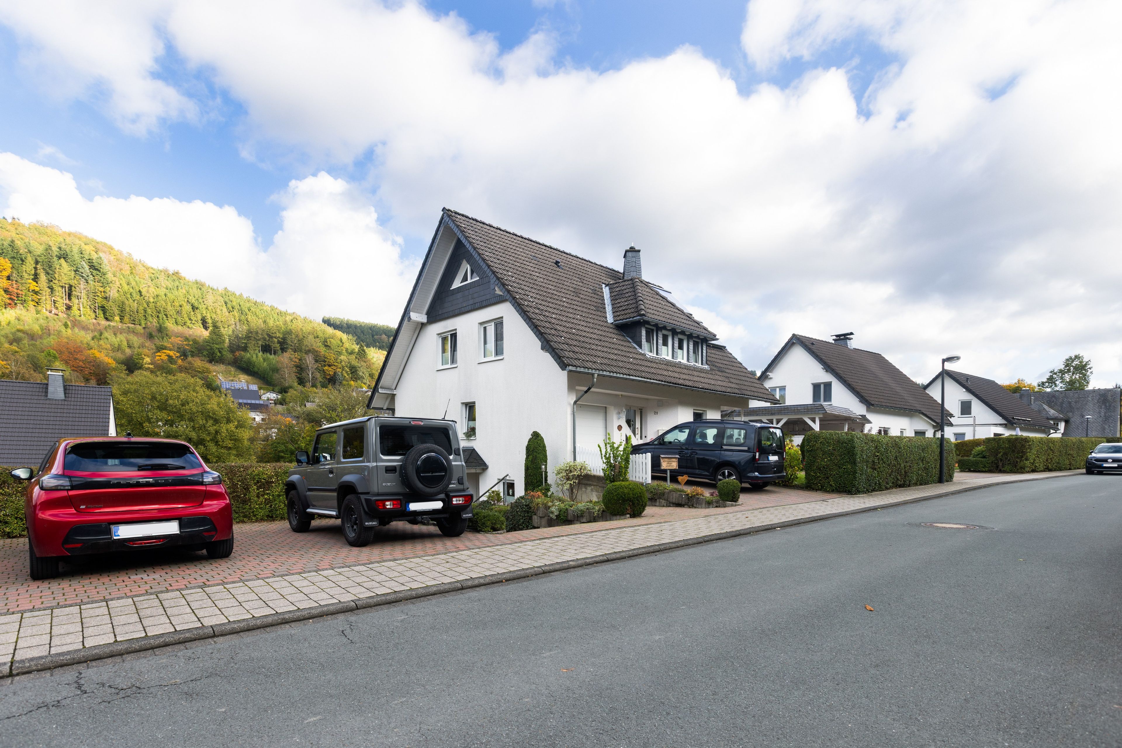 A residential street with houses, parked cars, and a hilly backdrop.
