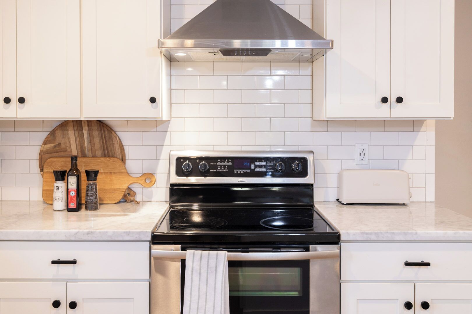 A kitchen with white cabinets, a stainless steel stove, a toaster, and a wooden cutting board with bottles of olive oil and salt.