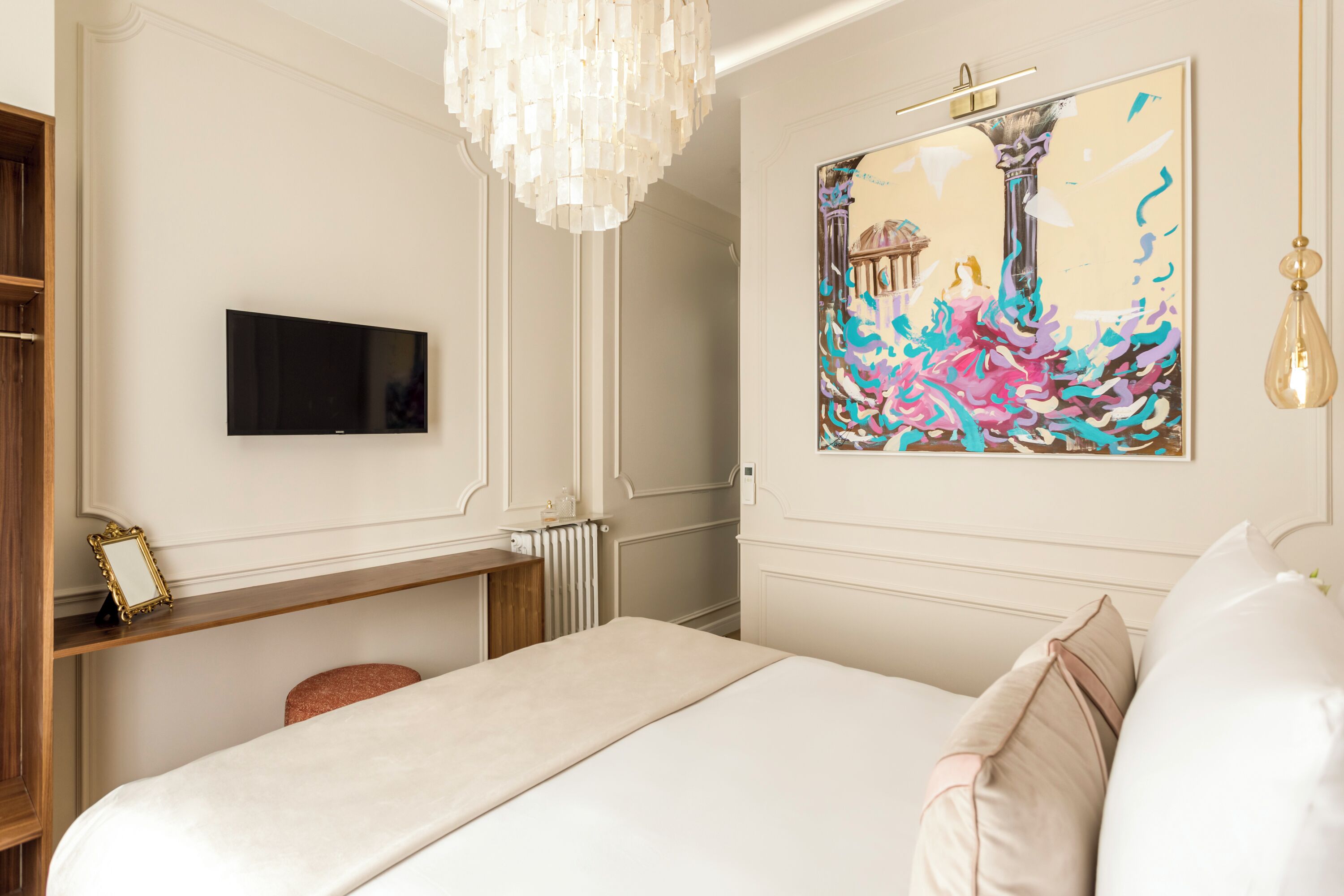 A hotel room with a bed, a television, a painting, and a chandelier.