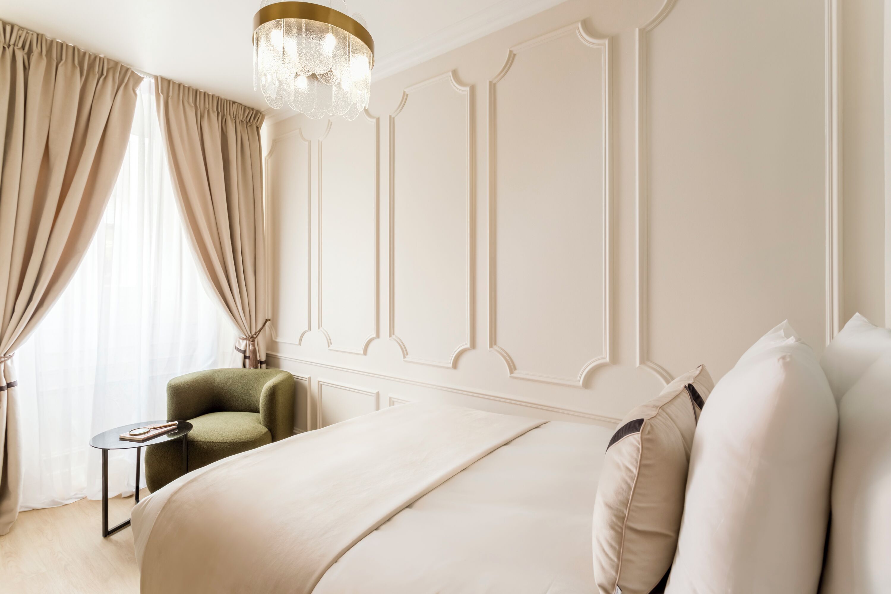 A hotel room with a large bed, a green armchair, a small table, and a chandelier.