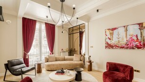 A modern living room with a beige sofa, a black chair, and two red velvet chairs. There is a round coffee table with books and a small lamp. The room has large windows with red curtains, a chandelier, and a painting on the wall. - Merveil Signature Suite - Kleber (Paris)