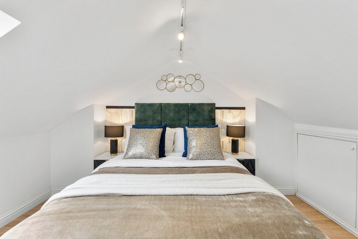 Une chambre à coucher avec un grand lit, deux tables de chevet, une tête de lit recouverte d’un tissu vert et un luminaire fixé au plafond.