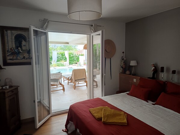 A bedroom with a bed, a red bedspread, a yellow towel, a painting, a hat, and a view of an outdoor area with a pool and greenery.