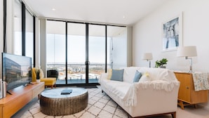 Un salón moderno con un sofá blanco, una mesa redonda de centro y un televisor sobre un mueble de madera. Amplias ventanas ofrecen vistas a la ciudad.