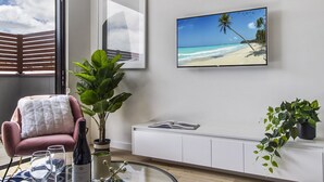 A modern living room with a glass coffee table, a pink armchair, a large potted plant, a flat-screen TV on the wall, and a view of the beach through a window. - Boutique Apartment in Quiet, Sought-after Suburb (Melbourne)