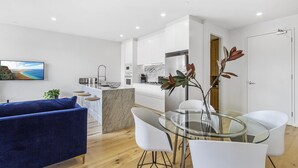 A modern living room with a blue sofa, a glass dining table with white chairs, a kitchen area with white cabinets, and a large flat-screen TV on the wall. - Boutique Apartment in Quiet, Sought-after Suburb (Melbourne)