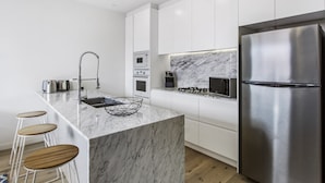 A modern kitchen with white cabinets, a stainless steel refrigerator, a marble countertop, and wooden bar stools. - Boutique Apartment in Quiet, Sought-after Suburb (Melbourne)