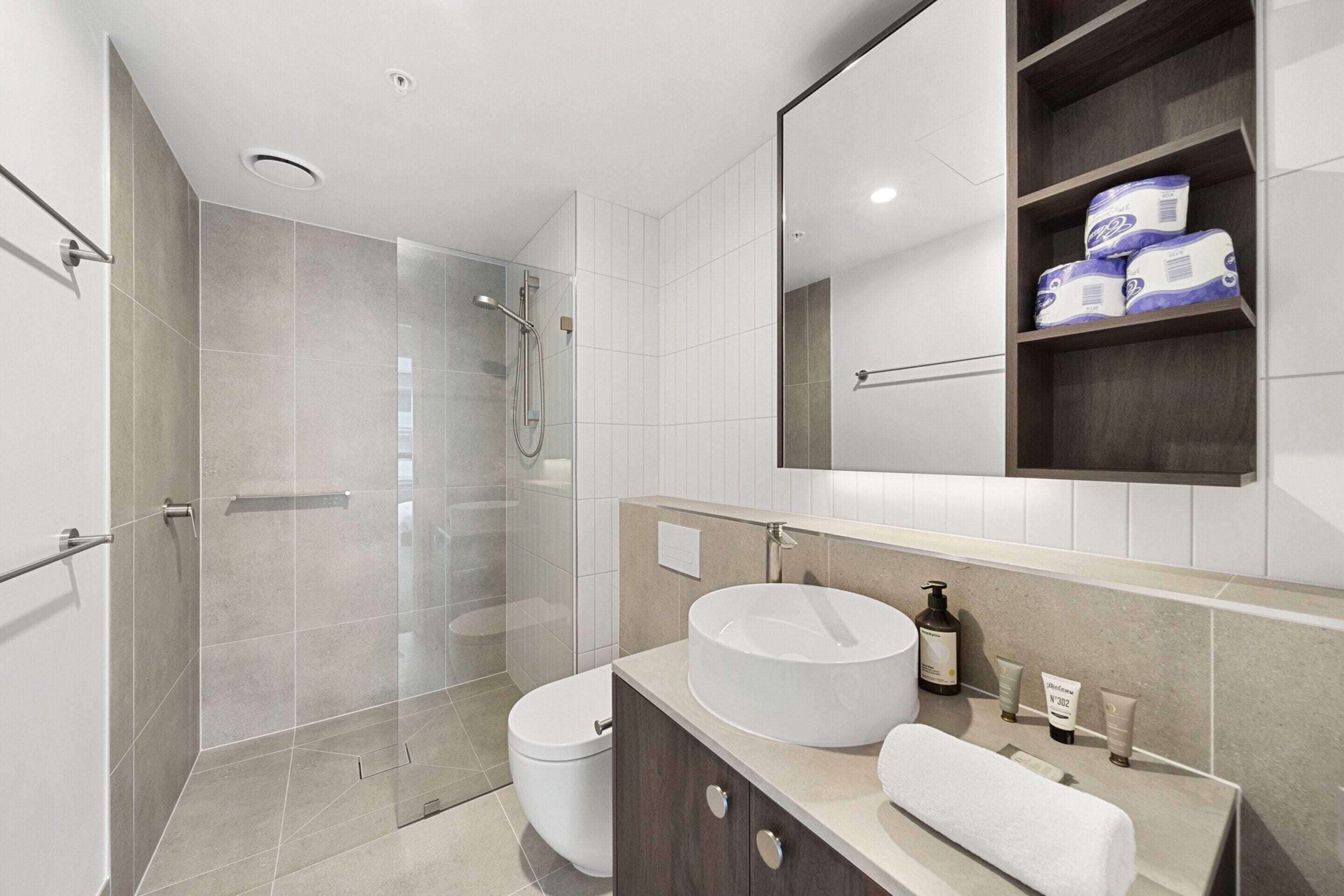 Un baño moderno con ducha envuelta en vidrio, un lavamanos redondo y un mueble de tocador con artículos de aseo.
