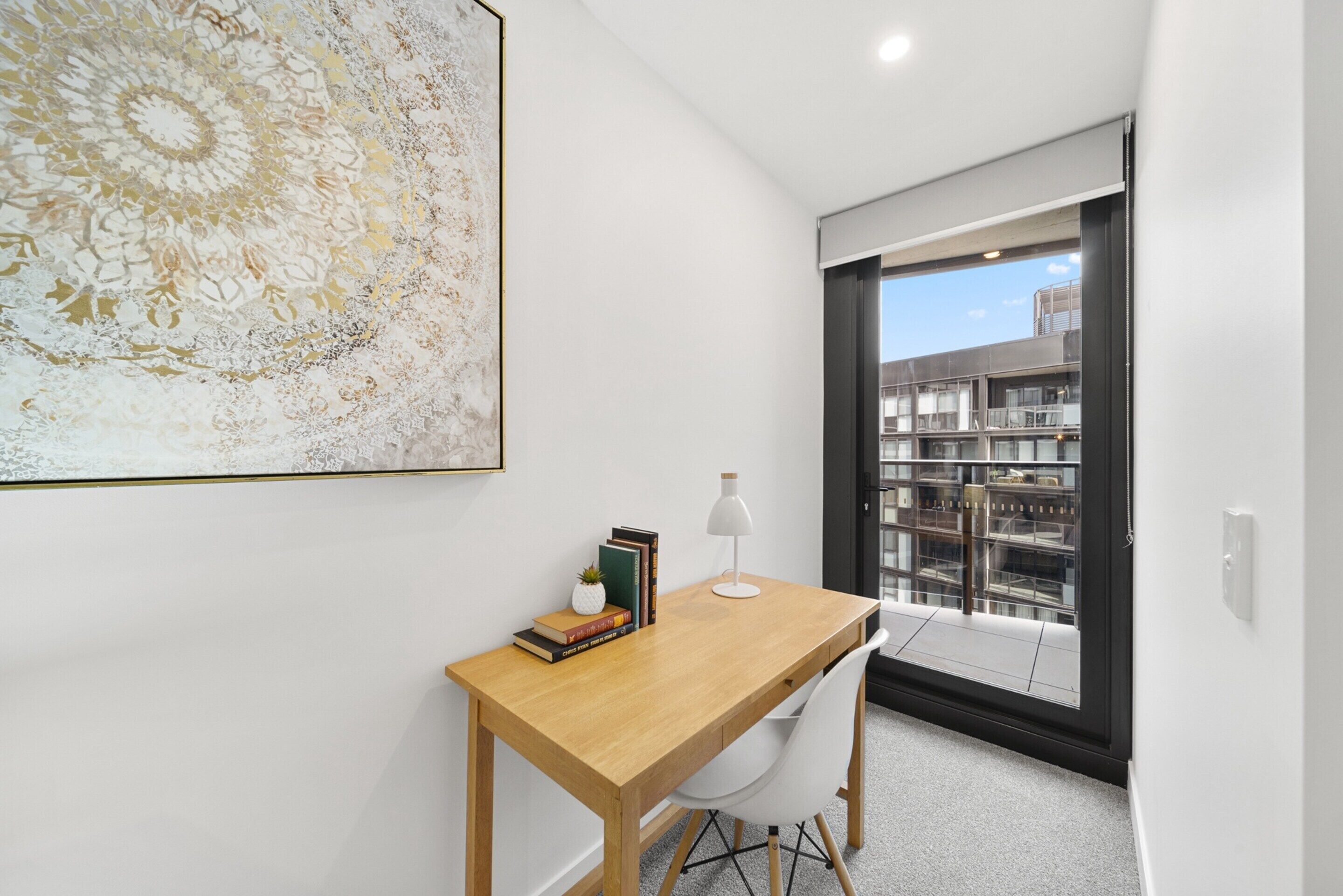 Ein modernes Zimmer mit einem Holzbüro, einem weißen Stuhl, einem großen abstrakten Gemälde und Blick auf Gebäude durch eine Glastür.