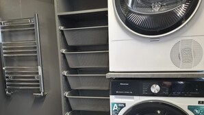 A stacked washer and dryer unit with a metal rack and a metal towel rack against a grey wall.