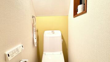 A small, narrow bathroom with a toilet, a hand dryer, and a small shelf with toiletries.