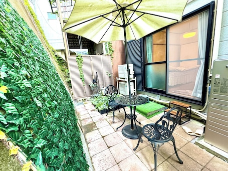 A patio area with a table and chairs, a large umbrella, and a view of a building with a window.