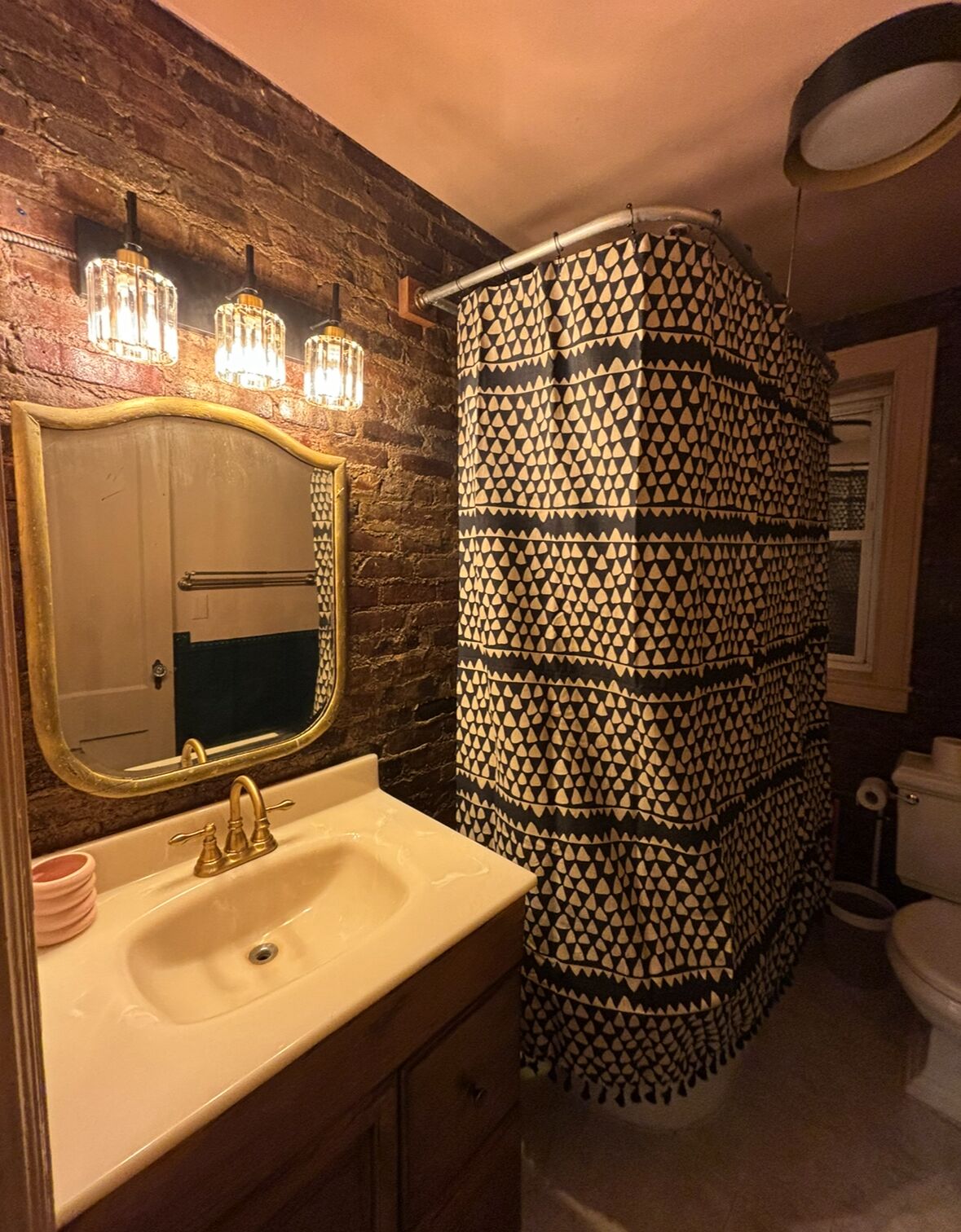 A bathroom with a stone wall, a gold-framed mirror, a sink with a gold faucet, and a patterned shower curtain.