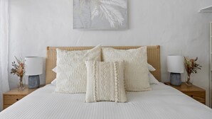A neatly made bed with white bedding and decorative pillows, flanked by two bedside tables with lamps and vases of dried flowers. - Chic 2-bed Cabin-style Home by Mooloolaba Beach (Sunshine Coast)