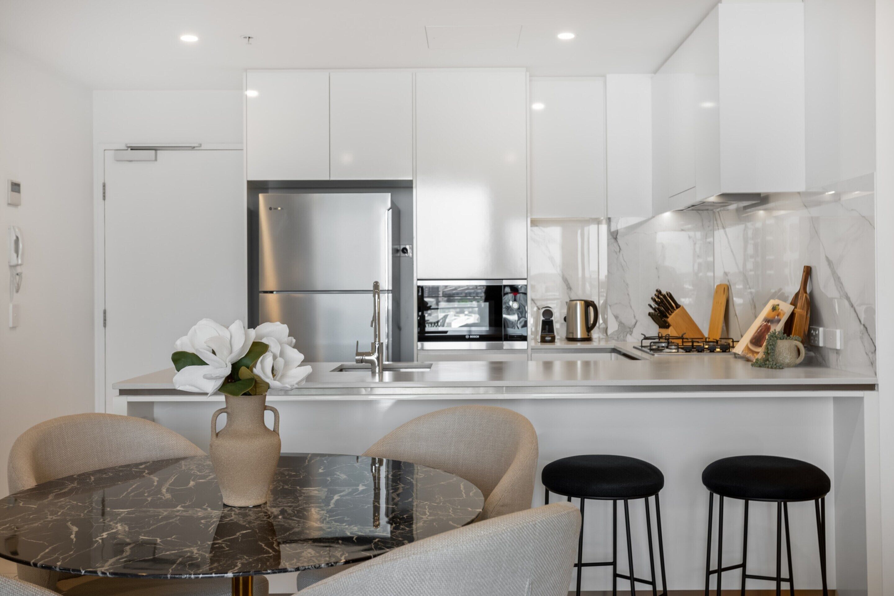 A modern kitchen with white cabinets, a stainless steel refrigerator, a marble countertop, and a dining area with a glass table and two bar stools.