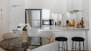 A modern kitchen with white cabinets, a stainless steel refrigerator, a marble countertop, and a dining area with a glass table and two bar stools. - Riverside 2-bed by Brisbane's Landmarks (Brisbane)