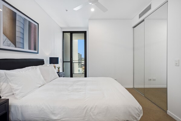 A modern hotel room with a large bed, a nightstand, a desk lamp, a ceiling fan, a large window, and a sliding door leading to a balcony.