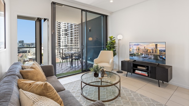 A modern living room with a sofa, a glass coffee table, a TV, and a sliding glass door leading to an outdoor patio with a city view. - Riverside 2-bed by Brisbane's Landmarks (Brisbane)