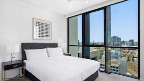 A modern hotel room with a large bed, a nightstand, a framed picture on the wall, and a city view through large windows. - Riverside 2-bed by Brisbane's Landmarks (Brisbane)