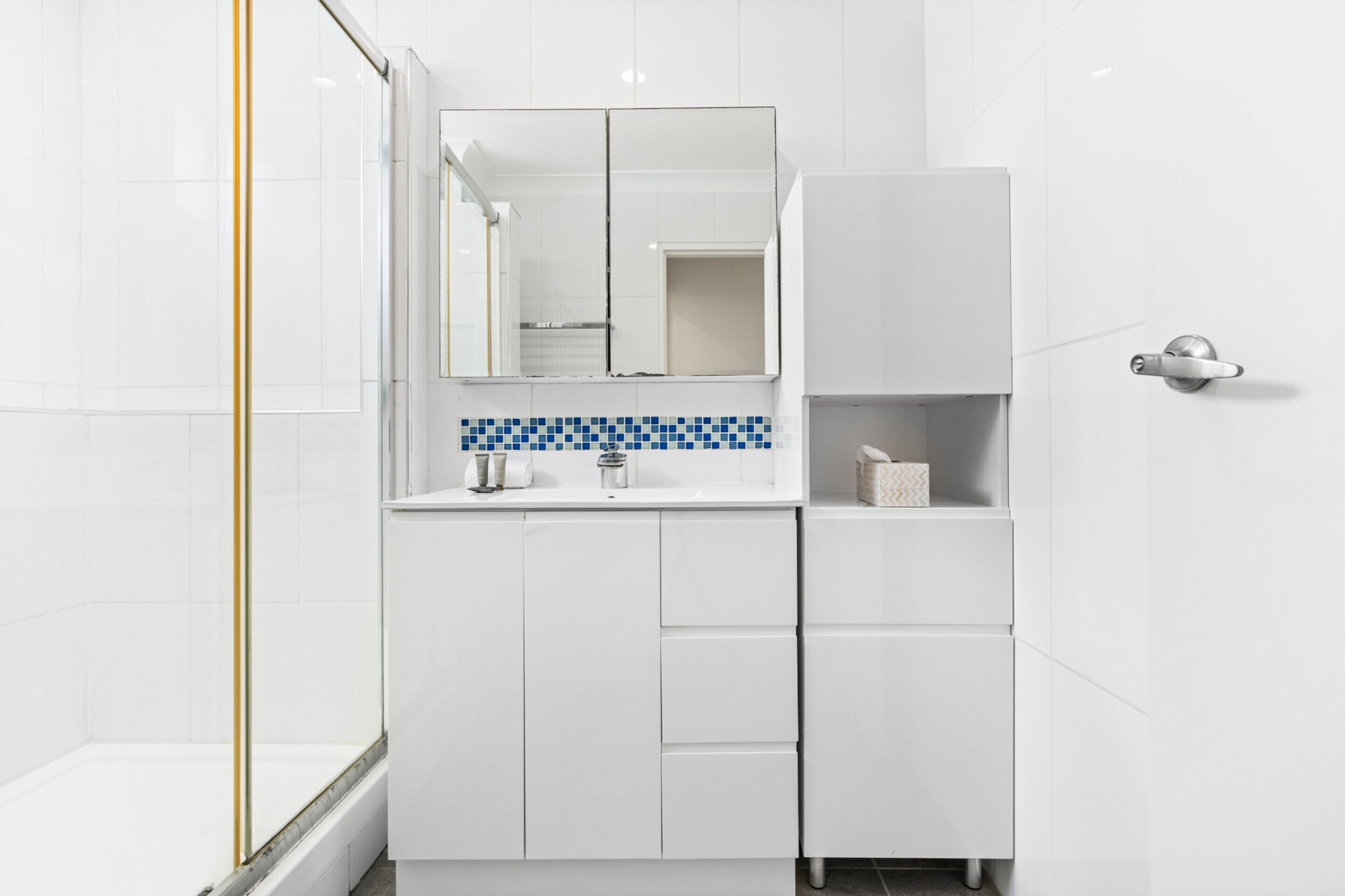 A modern bathroom with white cabinetry, a blue-tiled backsplash, and a glass shower enclosure.