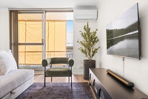 A modern living room with a sofa, a chair, a large window with a view, a flat-screen TV, and a potted plant. - Elegant 2-bed With Balcony by Parks and Amenities (Brisbane)