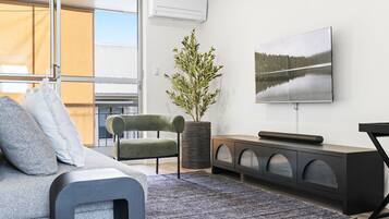 A modern living room with a grey sofa, a green armchair, a large flat-screen TV, a potted plant, and a black console table.