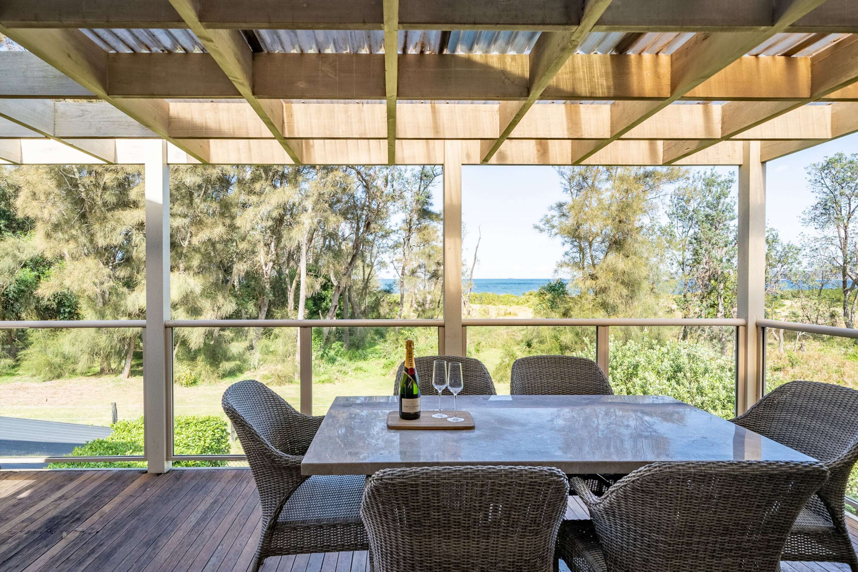 A covered outdoor dining area with wicker chairs and a table, overlooking a scenic landscape with trees and a view of the sea.