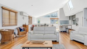 Un vaste espace de vie avec un canapé blanc, des fauteuils en osier et une table basse en bois. On aperçoit, au fond, une cuisine avec des meubles blancs et une lucarne.
