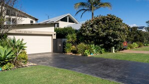 Une maison moderne à deux étages, dotée d’un garage, entourée de pelouses bien entretenues et de plantes tropicales.