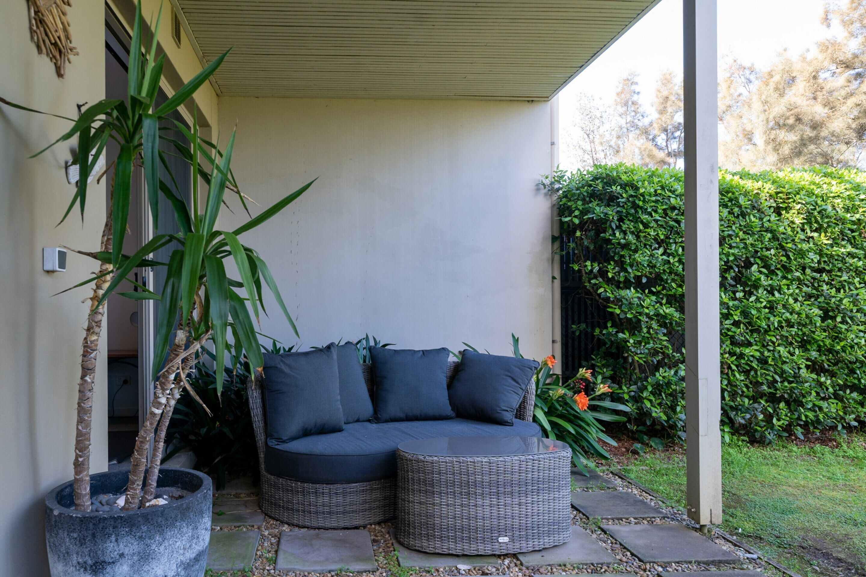 A corner seating area with a dark-colored sofa and a round wicker table, a potted plant, and a hedge in the background.