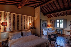 A cozy bedroom with a large bed, a smaller bed, and a chair. There is a window with a view of trees and a lamp on the wall.
