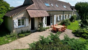 Une maison de campagne avec un toit en tuiles, une cheminée et une terrasse équipée d’une table et de chaises.