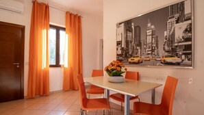 Un coin repas comprenant une table, quatre chaises orange, un vase de fleurs et un tableau représentant un paysage urbain accroché au mur.