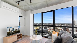 A modern living room with a large window, a flat-screen TV, a grey sofa, and a wooden entertainment unit. - Stylish 1-bed With Pool Gym & Stunning Views (Melbourne)