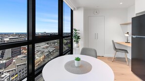 A modern room with a large window, a round table with a potted plant, a chair, a desk with a lamp, and a refrigerator. - Stylish 1-bed With Pool Gym & Stunning Views (Melbourne)