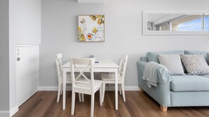 Una sala moderna con una mesa de comedor blanca y sillas, un sofá azul claro con cojines estampados, y un ventanal que proporciona luz natural.
