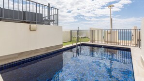 Una piscina en la azotea con vista al mar, rodeada por una valla metálica y un pequeño espacio cubierto de césped.