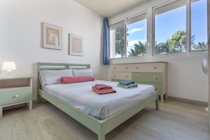 Ein Schlafzimmer mit Bett, Nachttisch, Kommode und großen Fenstern.
